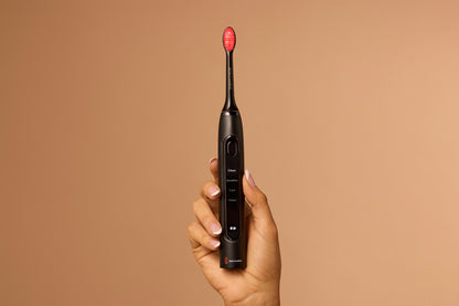 Hand holding a black electric toothbrush with a red bristle head against a brown background